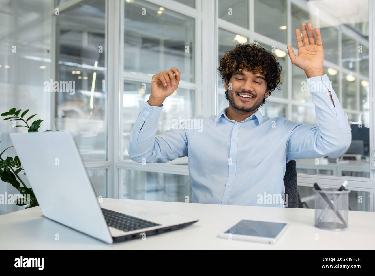 Curly islamic man in blue shirt raising arms for stretching back while sitting at workspace with closed eyes. Relaxed employee using free time for exe Stock Photo