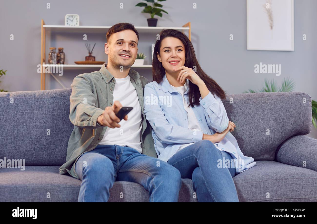 Cheerful couple is relaxing with remote control in their hands, choosing movie or show to watch. Stock Photo