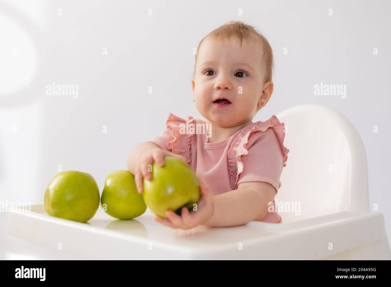 Cute baby in a baby chair nibbles apples on a white background. High quality photo Stock Photo