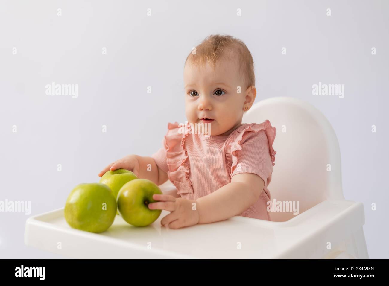 Cute baby in a baby chair nibbles apples on a white background. High quality photo Stock Photo