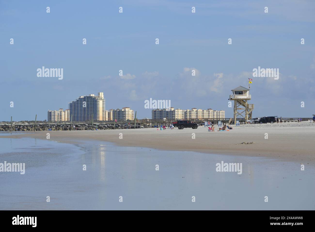 The backdrop of Ponce Inlet beach features a lifeguard station, parked cars, and people unwinding by the jetty, with the skyline of New Smyrna Beach. Stock Photo