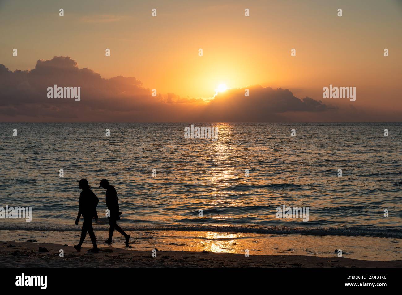 Two people walking on a beach at sunset. The sky is orange and the water is calm Stock Photo