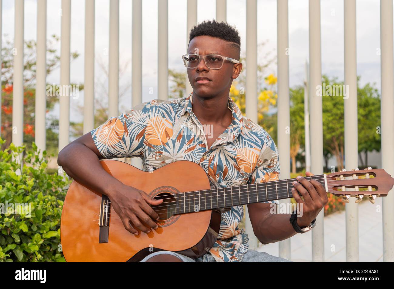 A young African American male musician plays his guitar, seated outdoors with a serene expression and surrounded by a lush urban environment. Stock Photo