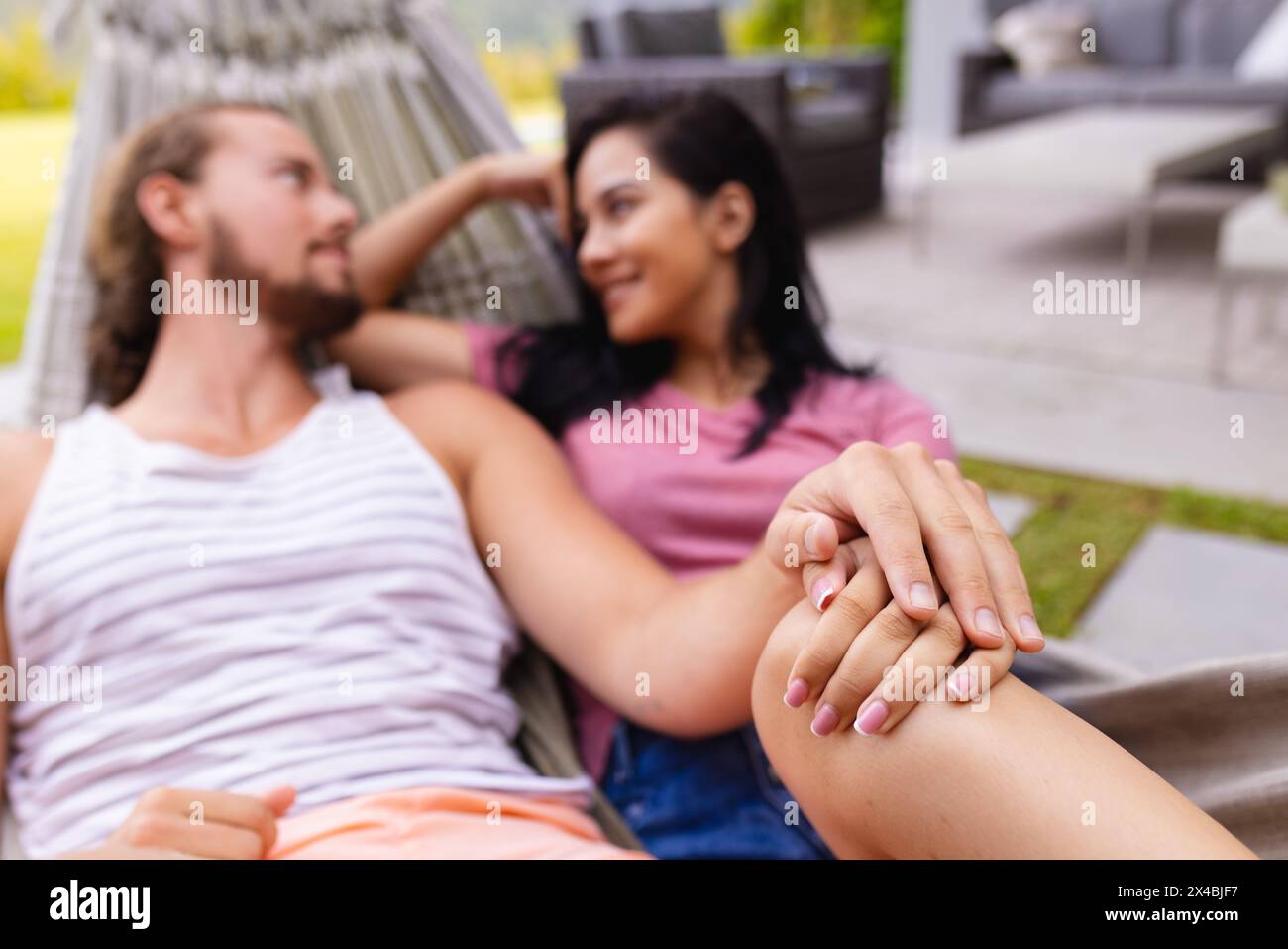 Diverse couple relaxing outdoors by pool, holding hands. Biracial woman with dark hair and Caucasian man with beard and long hair, sharing quiet momen Stock Photo