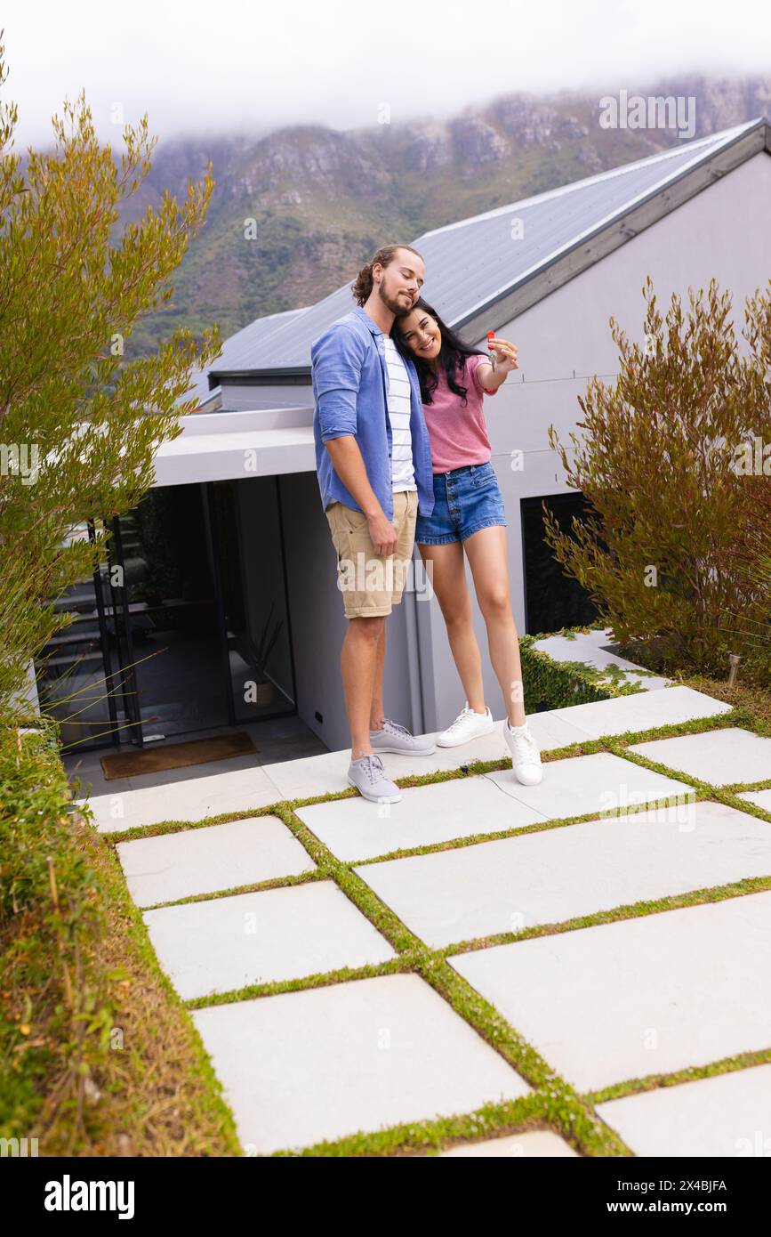 Diverse couple embracing outdoors, enjoying their new home. Biracial woman with long dark hair and Caucasian man with beard are smiling warmly, unalte Stock Photo