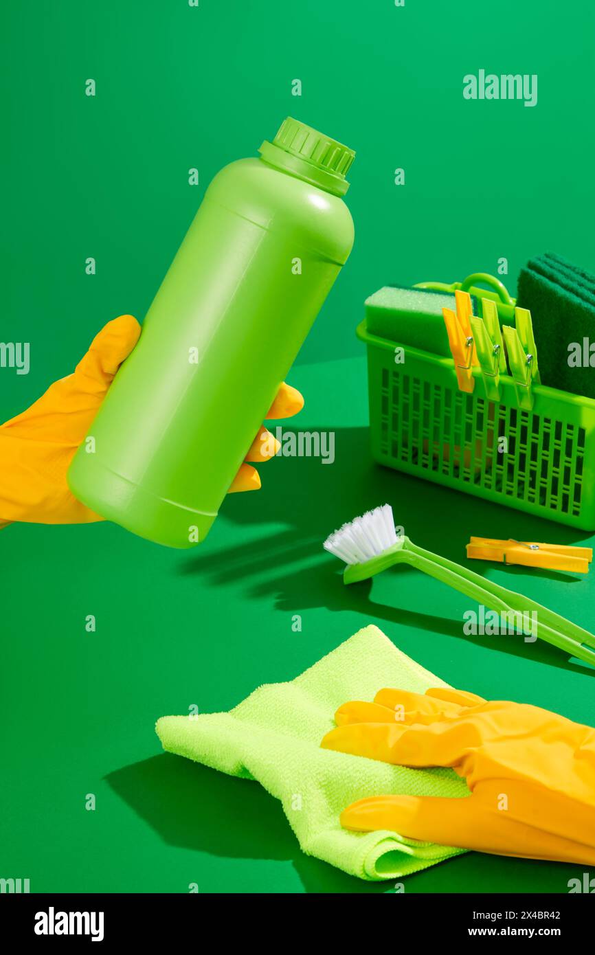 Cleaner with yellow rubber gloves holding unlabeled detergent bottle on green background. In the back is a plastic basket with cleaning supplies. Hous Stock Photo