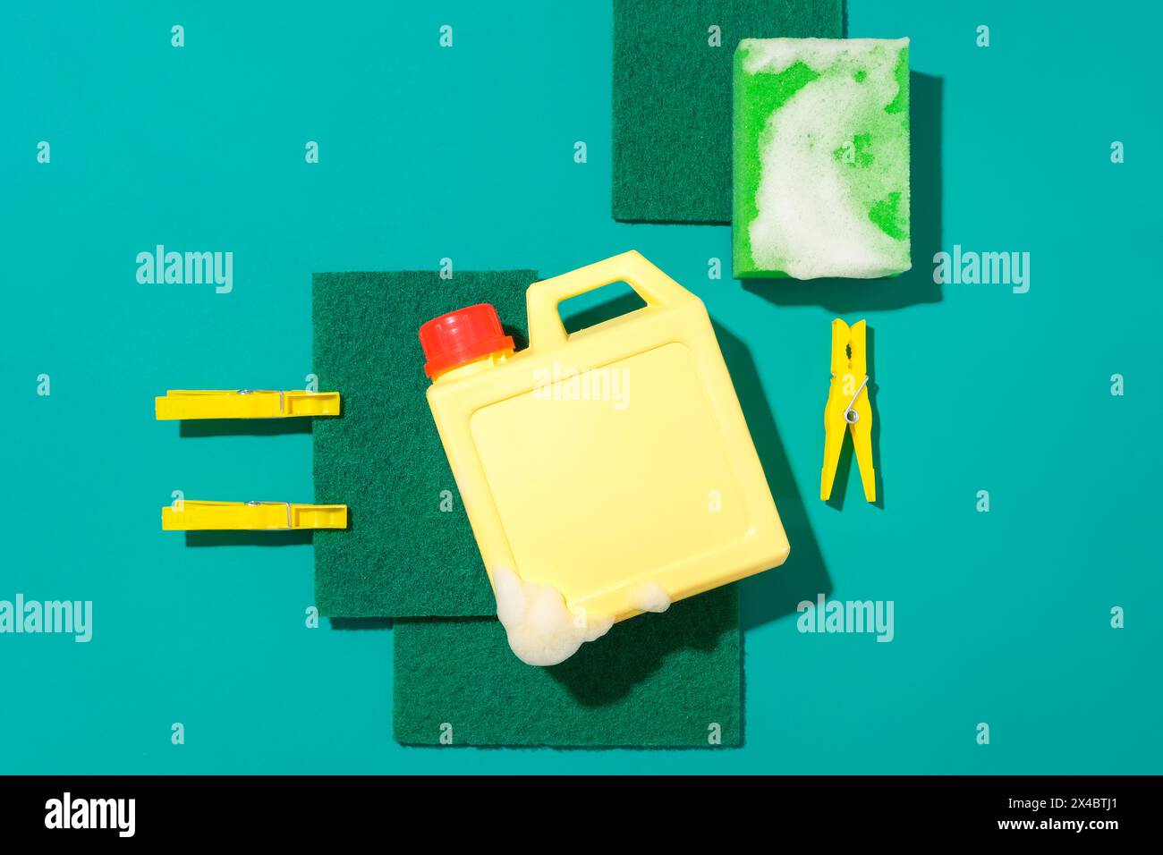 Close-up of an unlabeled detergent bottle next to a sponge and yellow clothespins on a turquoise background. Advertising space for cleaning products. Stock Photo