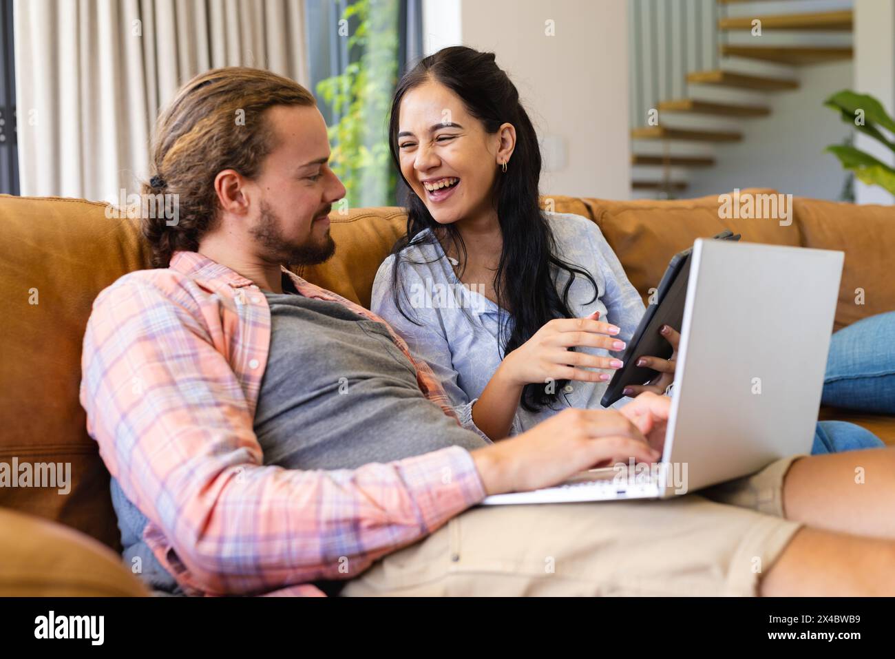 Biracial woman and Caucasian man, couple, laughing and looking at laptop at home. She has dark hair and he sports beard and long hair, both dressed ca Stock Photo