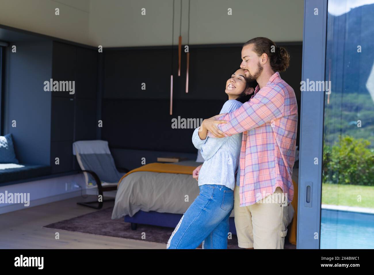 Diverse couple embracing in modern bedroom at home, copy space. She has curly brown hair, he has a beard and long hair, unaltered Stock Photo