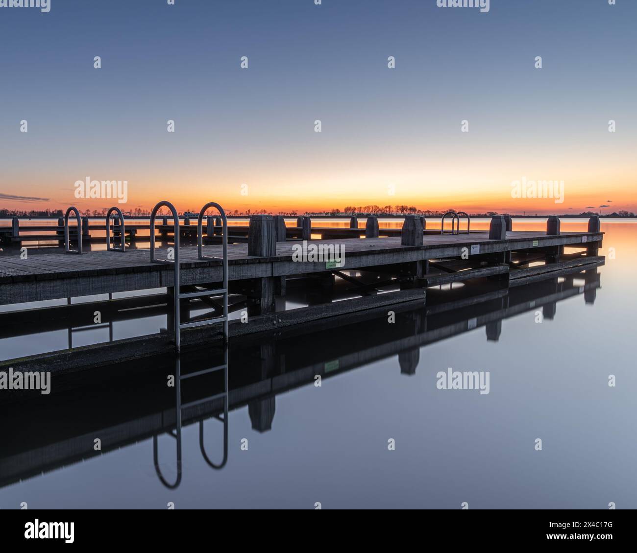 A calm sunrise in the recreational area De Hoorne on the Alkmaardermeer creates a pleasant reflection of the swimming pontoon. The sun is about to ris Stock Photo