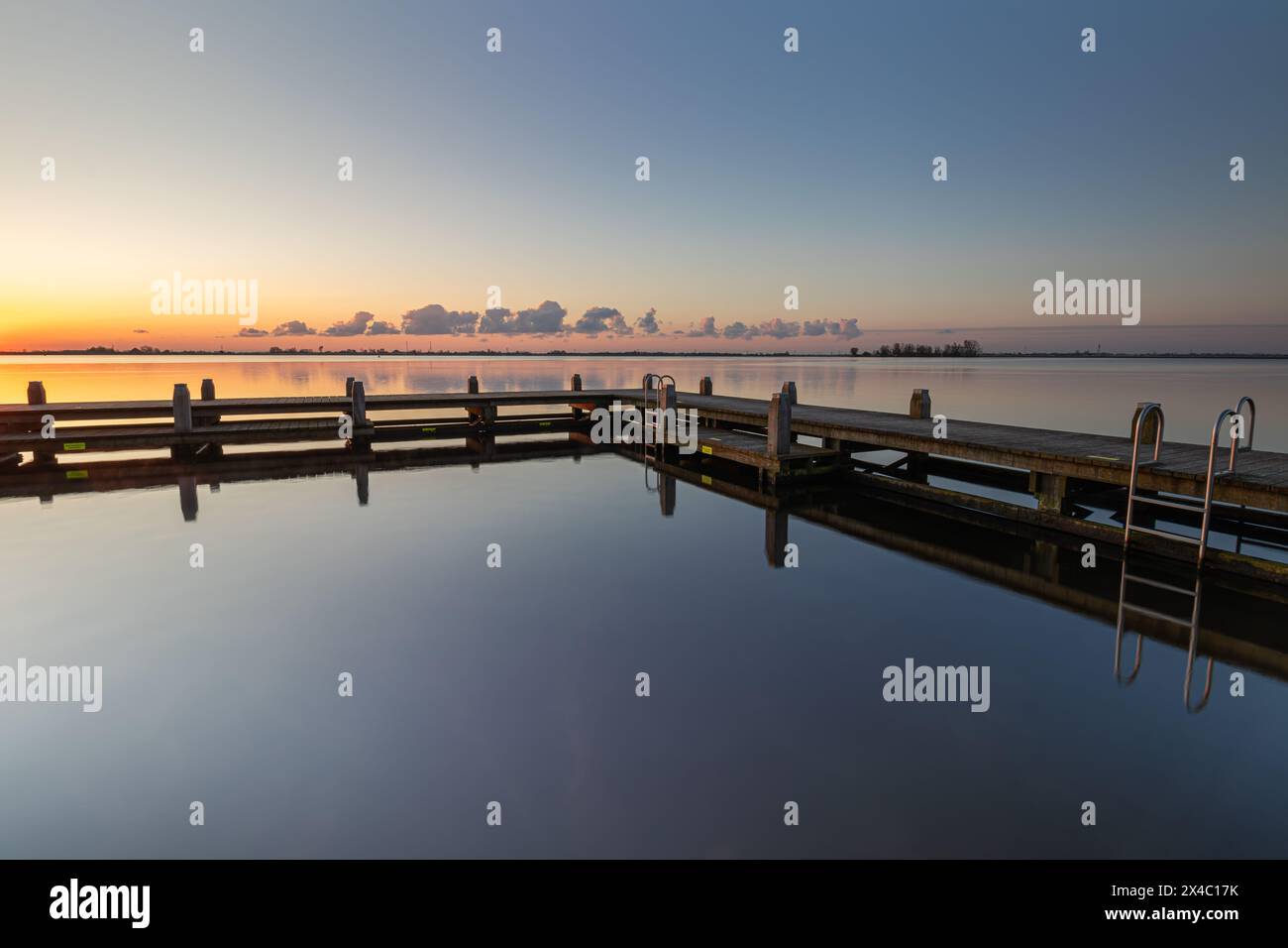 A calm sunrise in the recreational area De Hoorne on the Alkmaardermeer creates a pleasant reflection of the swimming pontoon. The sun is about to ris Stock Photo