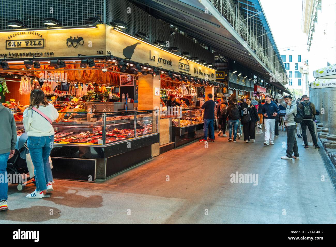 Ein beleuchteter Marktstand bietet eine reiche Auswahl an Schinken und Fleischwaren, Kunden im Hintergrund, Mercado de la Boqueria, berühmter Markt an Stock Photo