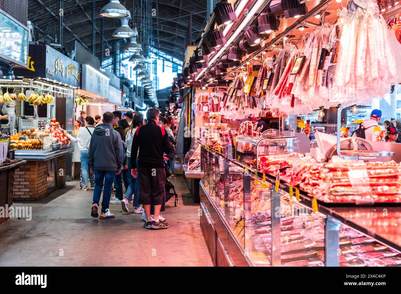 Ein beleuchteter Marktstand bietet eine reiche Auswahl an Schinken und Fleischwaren, Kunden im Hintergrund, Mercado de la Boqueria, berühmter Markt an Stock Photo