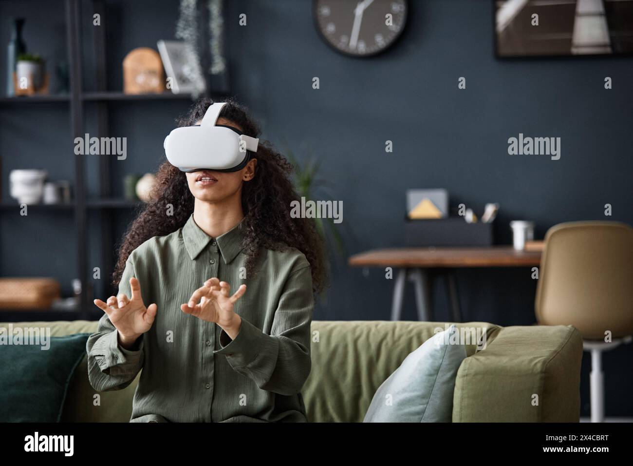 Young woman wearing VR headset sitting on sofa in living room typing text in the air, copy space Stock Photo