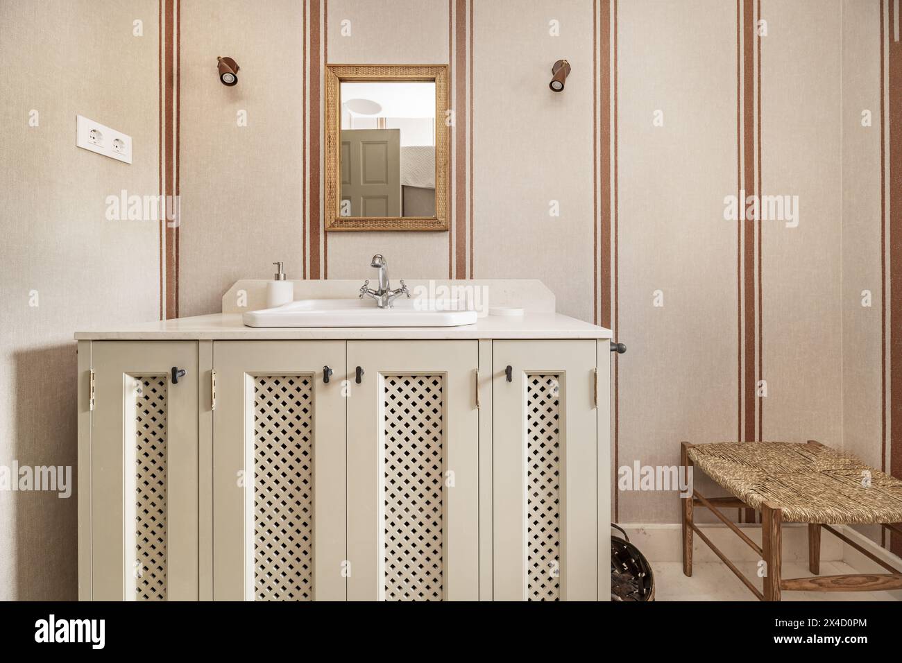 Bathroom with cream marble countertop, gold-framed mirrors, striped wallpaper, white porcelain sink on cabinet with wooden lattice doors Stock Photo
