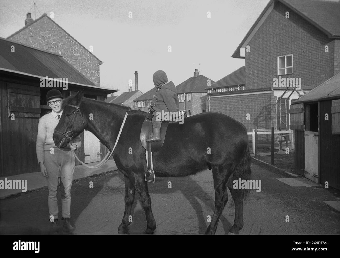 1960s, historical, outside at a suburban riding school, an infant child sitting on a horse by the stables, the horse's head being held a teenage girl, England, UK. Stock Photo