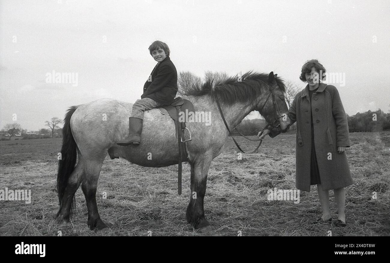 1960s, historical, outside at a suburban riding school, a young girl sitting backwards on a horse, hands on head, her mother holding the reins of the horse. Sitting this way on a horse helps in learning to balance when riding. Stock Photo