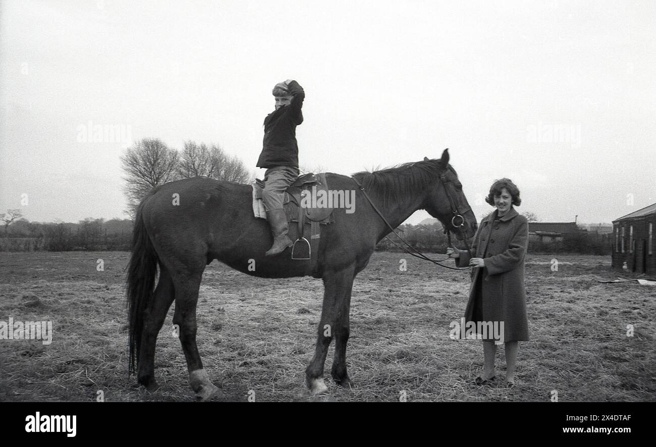 1960s, historical, outside at a suburban riding school, a young boy sitting backwards on a horse, hands on head, mother holding the reins of the horse. Sitting this way on a horse helps in learning to balance. Stock Photo