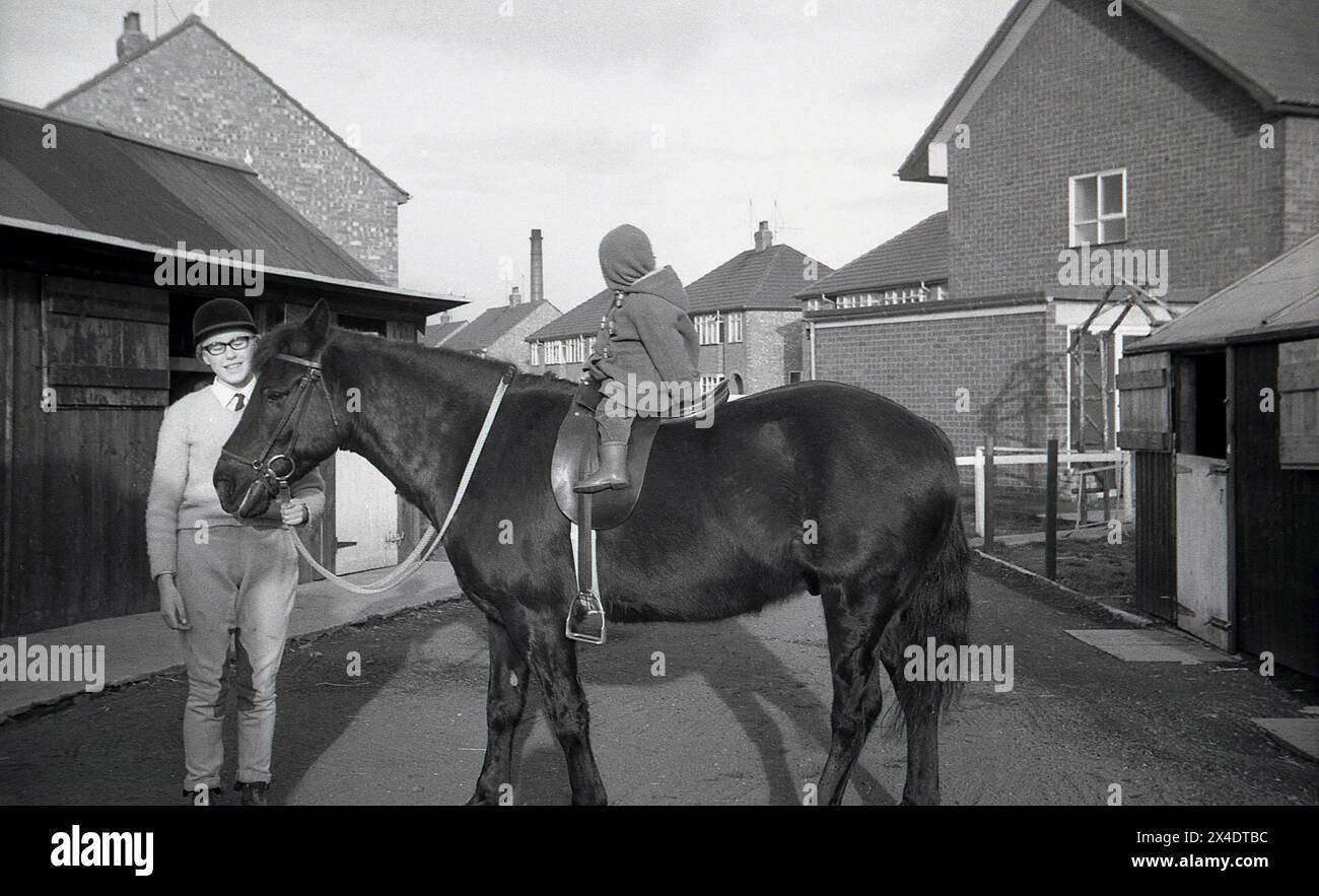 1960s, historical, outside at a suburban riding school, an infant child sitting on a horse by the stables, the horse's head being held a teenage girl, England, UK. Stock Photo