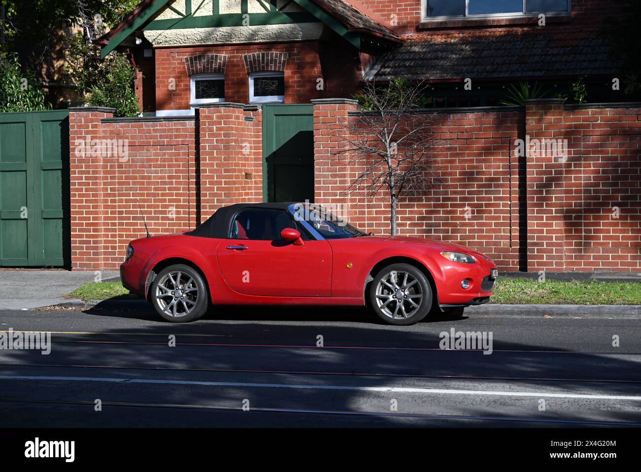 Side view of a bright red Mazda MX-5 convertible, parked in the street outside a suburban home with a red brick wall, during a sunny day Stock Photo