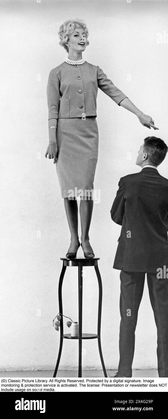 In the 1950s. A young woman seen standing on a piece of furniture higher than the man standing next to her. 1959 Stock Photo