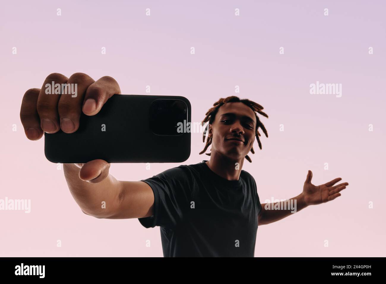 Dreadlocked young man holds a smartphone in a selfie pose against a pink studio backdrop. Gen Z man capturing a self-portrait. Stock Photo
