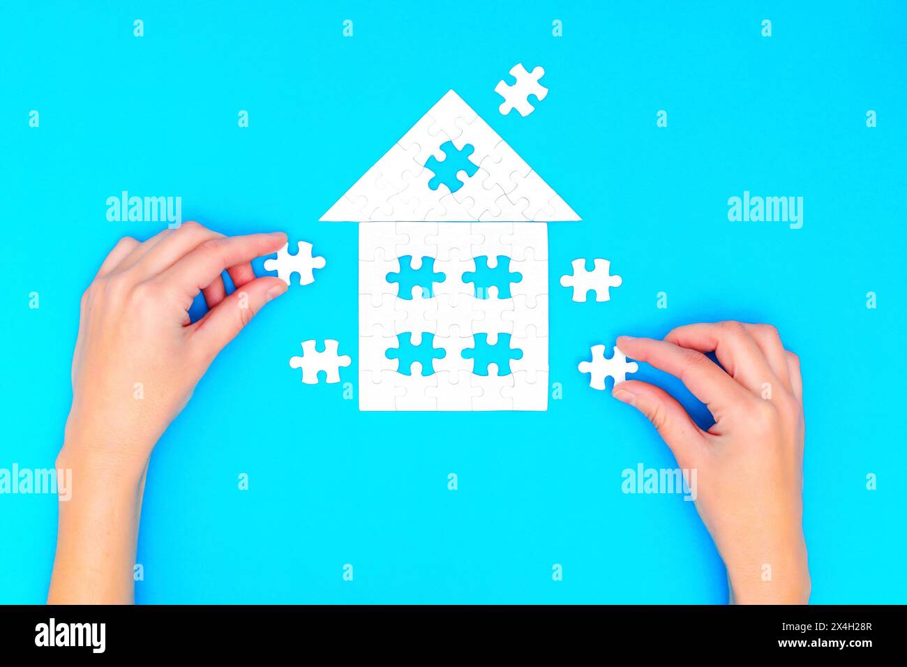 Hands assembling a puzzle in the shape of a house with a hip roof, set against a blue background. Stock Photo