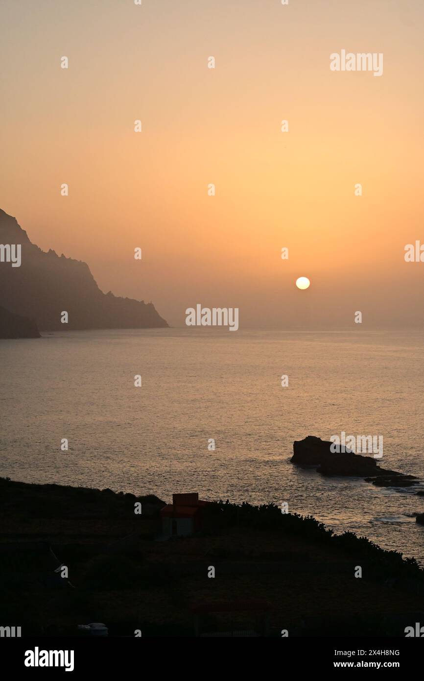 sunset over a calm sea with a distant shoreline, capturing the serene beauty of the sun setting against the backdrop of rugged cliffs and tranquil wat Stock Photo