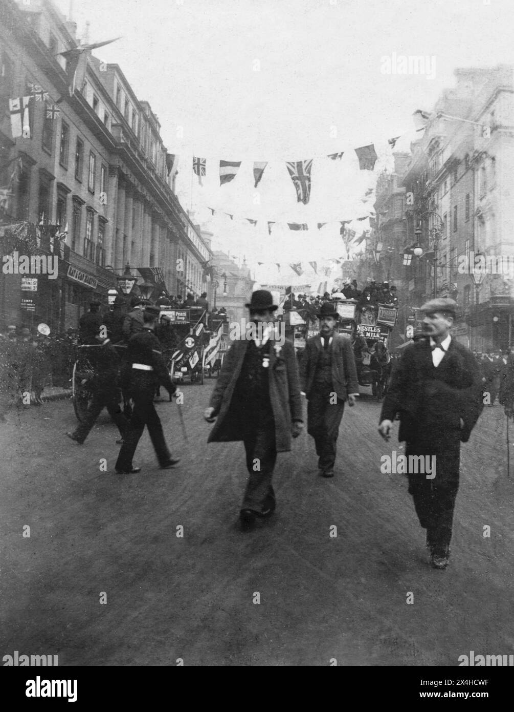 London. 1900 – A busy street scene in London. The street is decorated with flags and bunting for the procession held on 2 October 1900 to commemorate the return of The City of London Imperial Volunteers, who had served in South Africa during the Second Boer War. Horse-drawn omnibuses, a soldier and civilian pedestrians are on the road. A man in the foreground wears a bowler hat and a medal on his waistcoat. Stock Photo