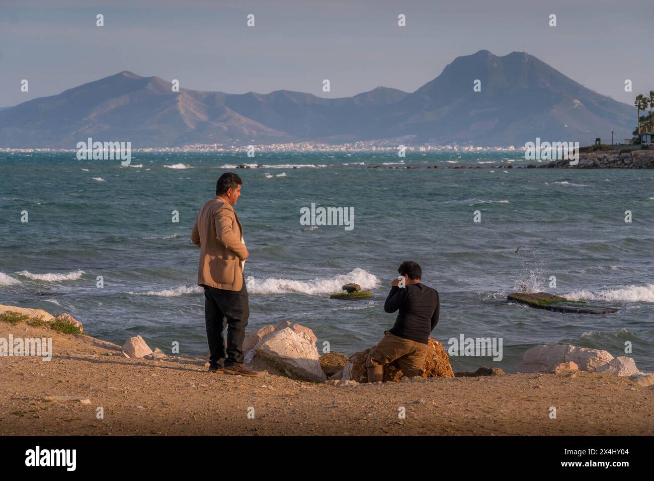 Two men talking on the coastline in Tunisia's Carthage district with the Boukornine mountains in the background during the hot summer day. Stock Photo