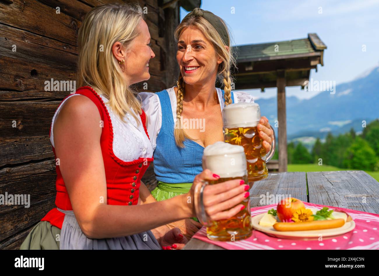 Happy girlfriends clinking beer mugs in front of the bavarian alpine mountains at a alpine cabin in traditional dirndls or tracht. Oktoberfest Bavaria Stock Photo