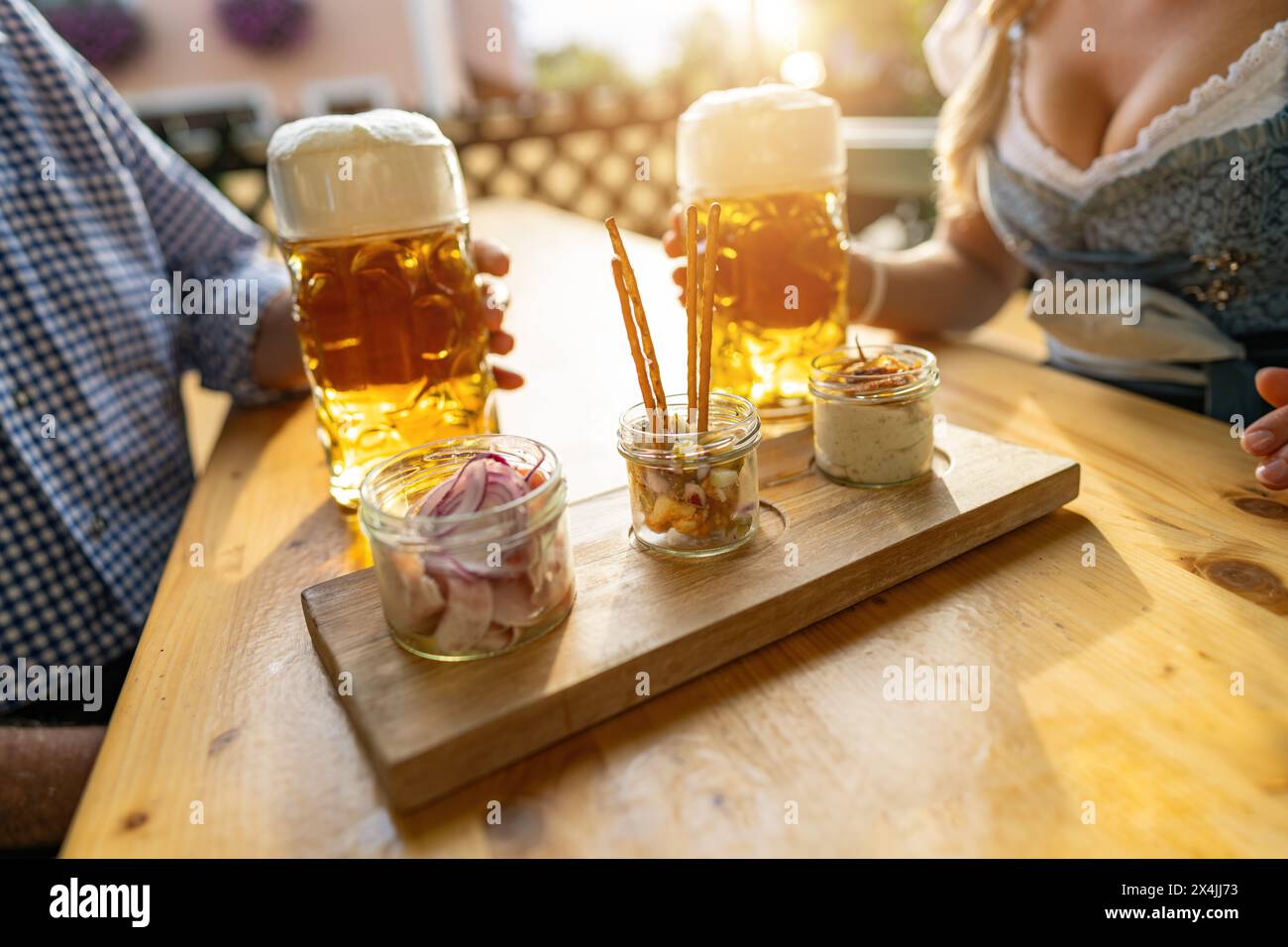 Bavarian Obatzda with pretzels and radishes and beer mugs, man and young woman in tracht in the background at beer garden or oktoberfest, Munich, Germ Stock Photo