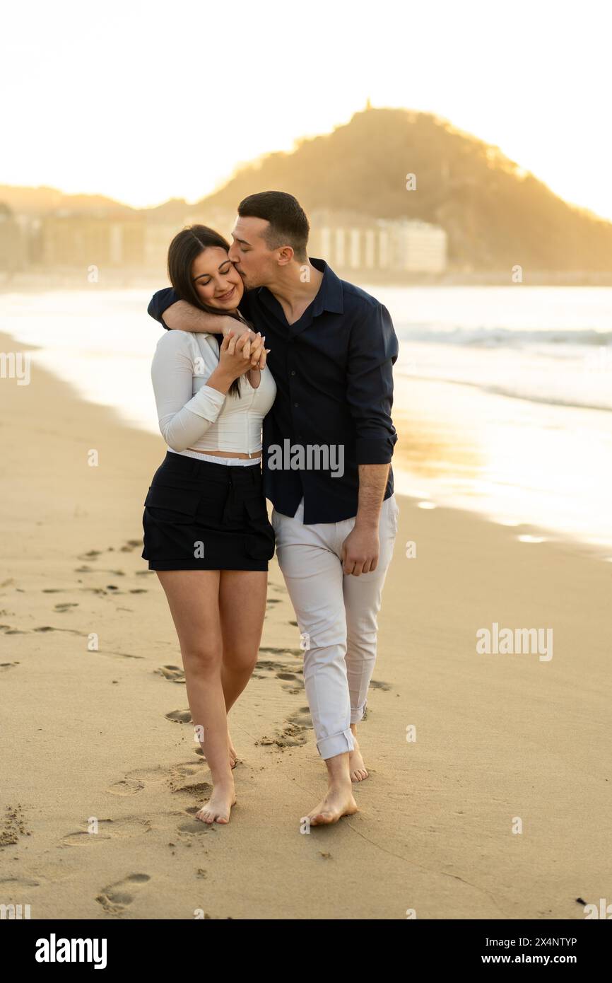 Vertical photo of a caucasian young couple kissing while walking barefoot along a beach during sunset Stock Photo