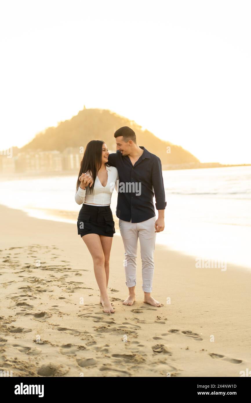 Vertical photo of a romantic Young couple walking embraced barefoot along a beach during sunset Stock Photo