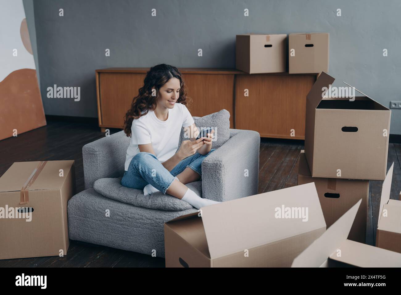 Casually dressed woman sitting comfortably in a chair using her smartphone, surrounded by boxes. Stock Photo