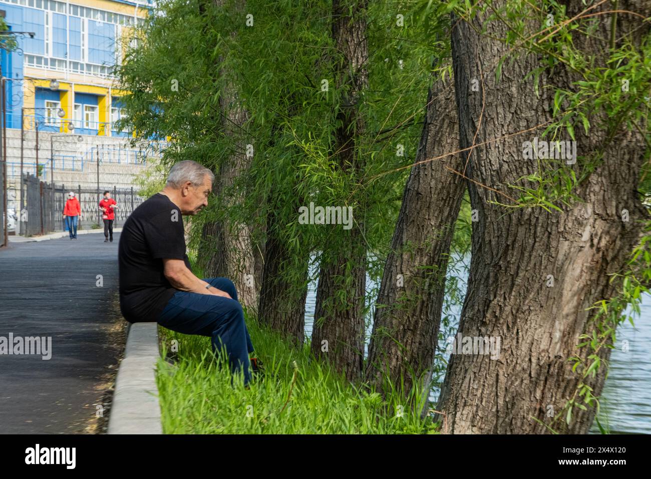 A man sits on a curb by the river, a woman jogs by the water Stock Photo