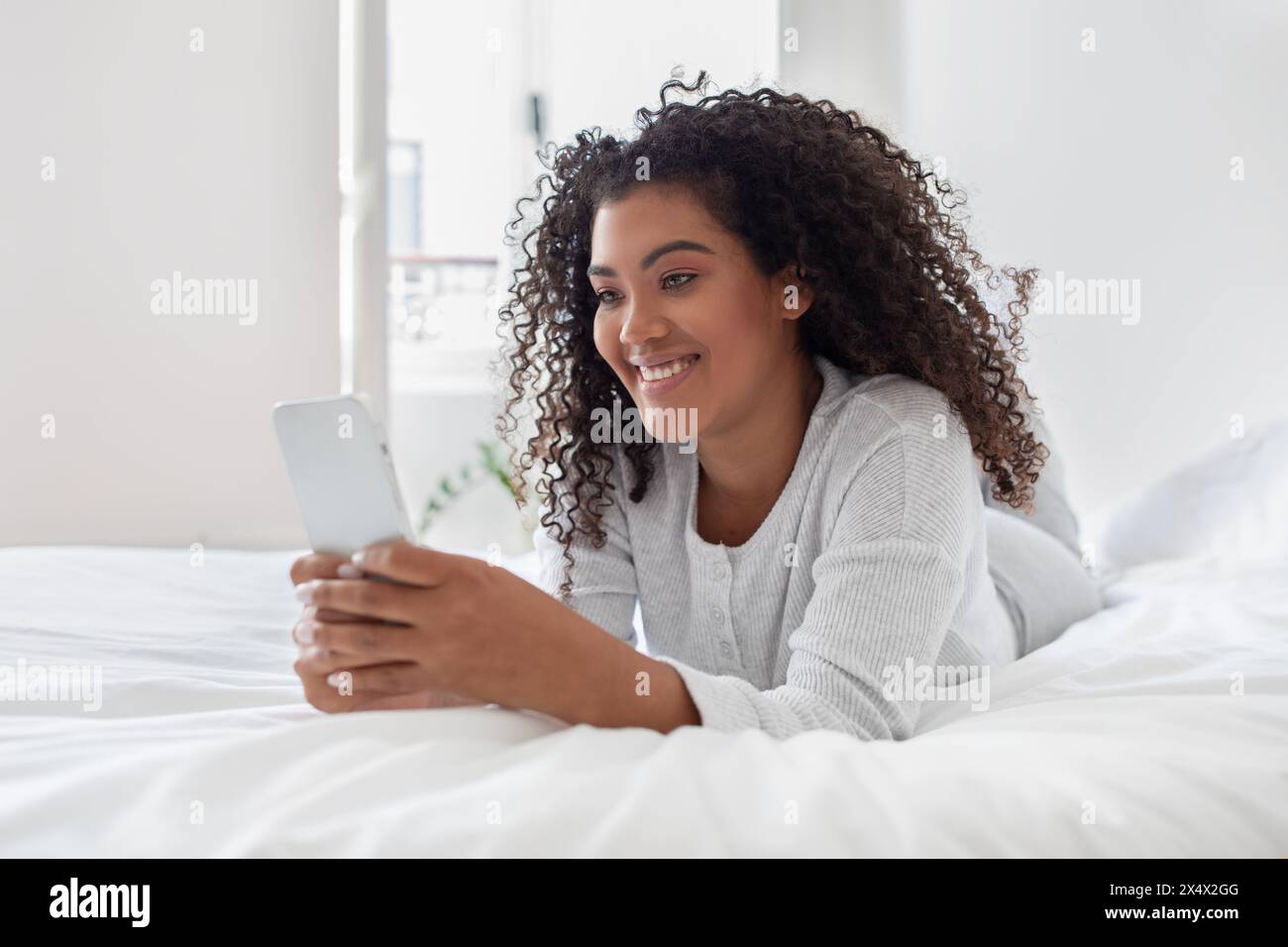 Young Woman Smiling While Using Smartphone in Bright Bedroom Setting Stock Photo