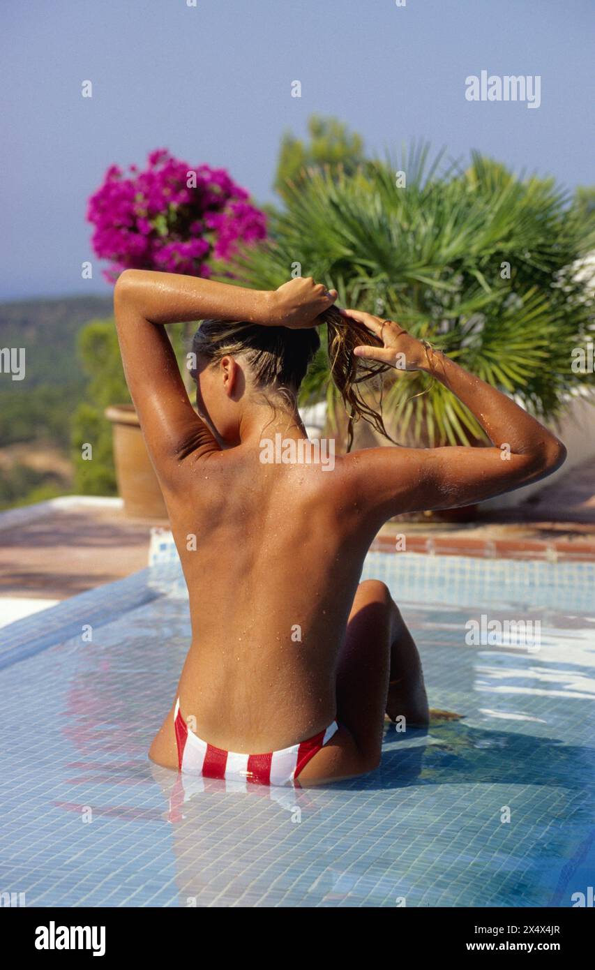 pretty young attractive woman nack from camera taking sun sit down on a pool green foliage background Stock Photo