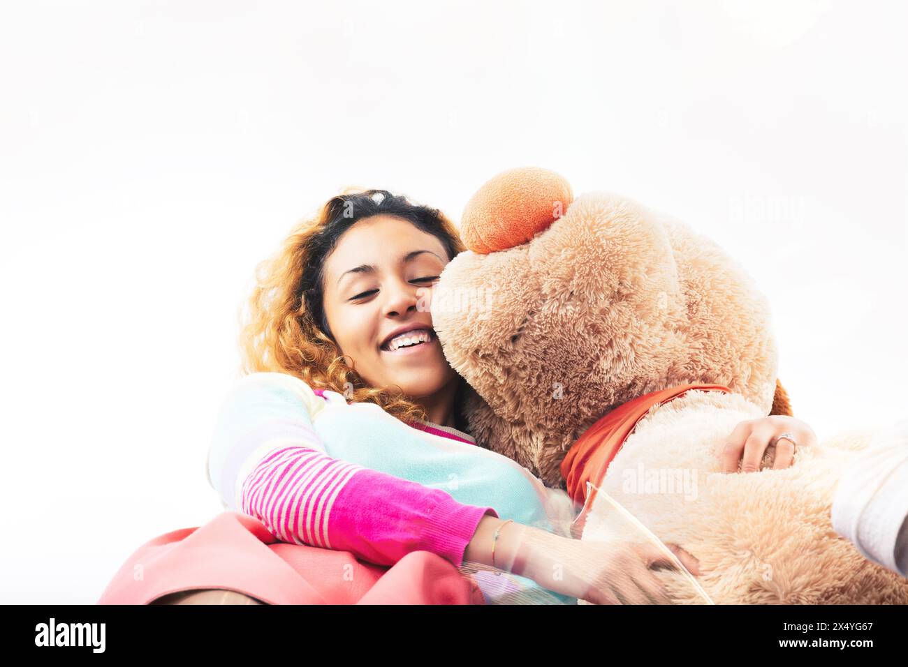Radiant young woman embraces a large teddy bear with a joyful smile, her colorful attire adding to the cheerful scene Stock Photo