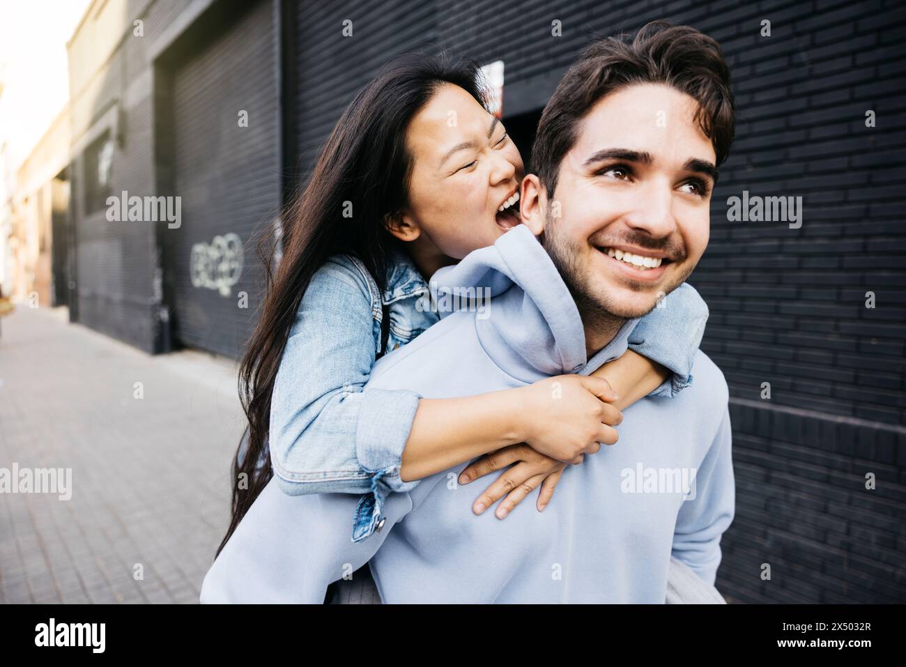 Young man carrying his girlfriend on his back while she tries to bite his ear, walking on the city street. Couple piggybacking on city street. Stock Photo