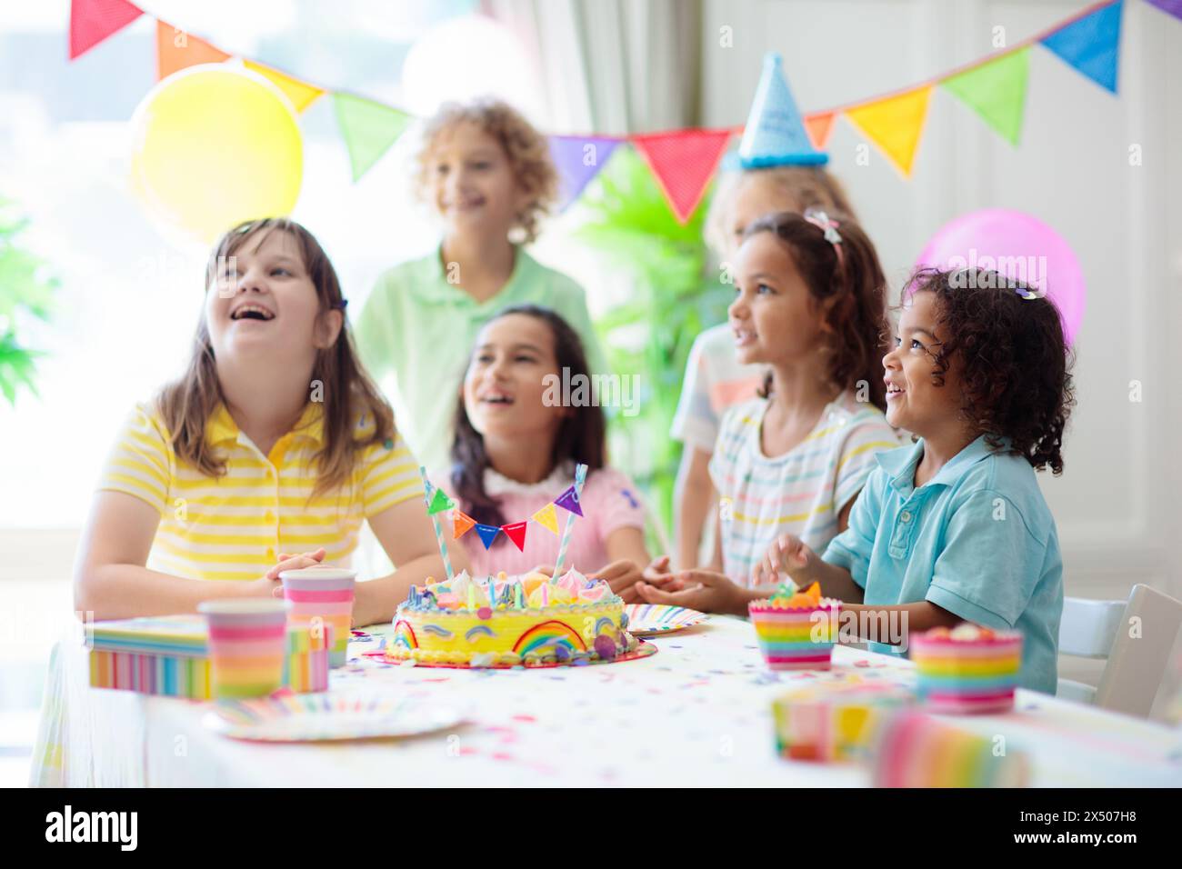 Kids birthday party. Children celebrate with colorful cake and gifts. Little curly boy blowing candles and opening birthday presents. Stock Photo