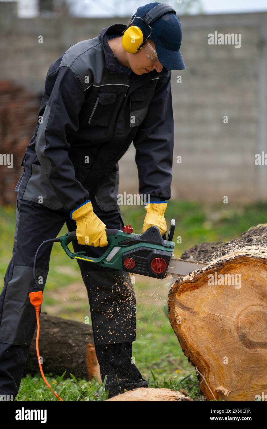 A man in uniform cuts an old tree in the yard with an electric saw Stock Photo