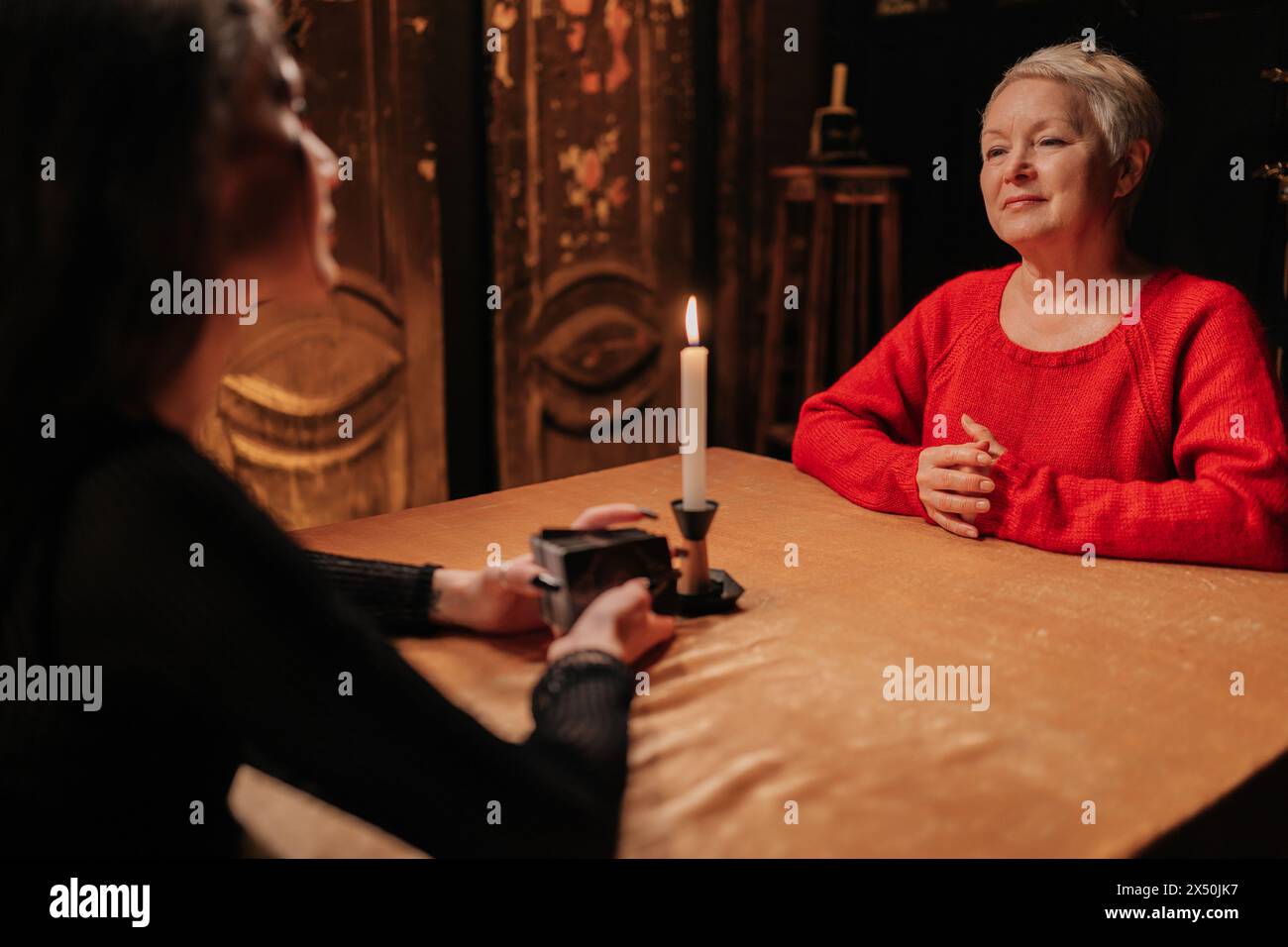 Rear view of enigmatic fortune teller reading tarot cards to mature adult woman client during spiritual ceremony in dark room by light of burning Stock Photo