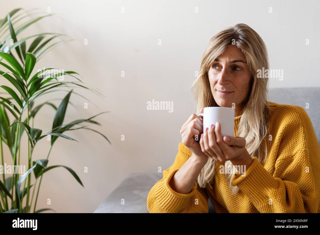 Mature woman in yellow sweater holding a white mug, thoughtfully looking away, with a houseplant beside her Stock Photo