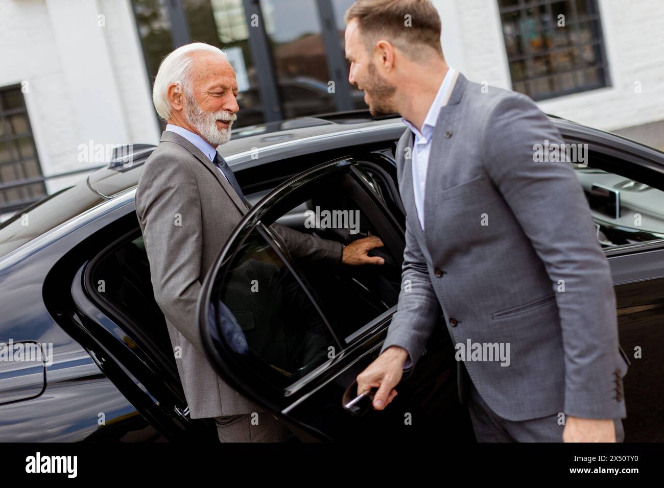 Well-dressed senior businessman is warmly greeted by his chauffeur as he approaches a sleek black car Stock Photo