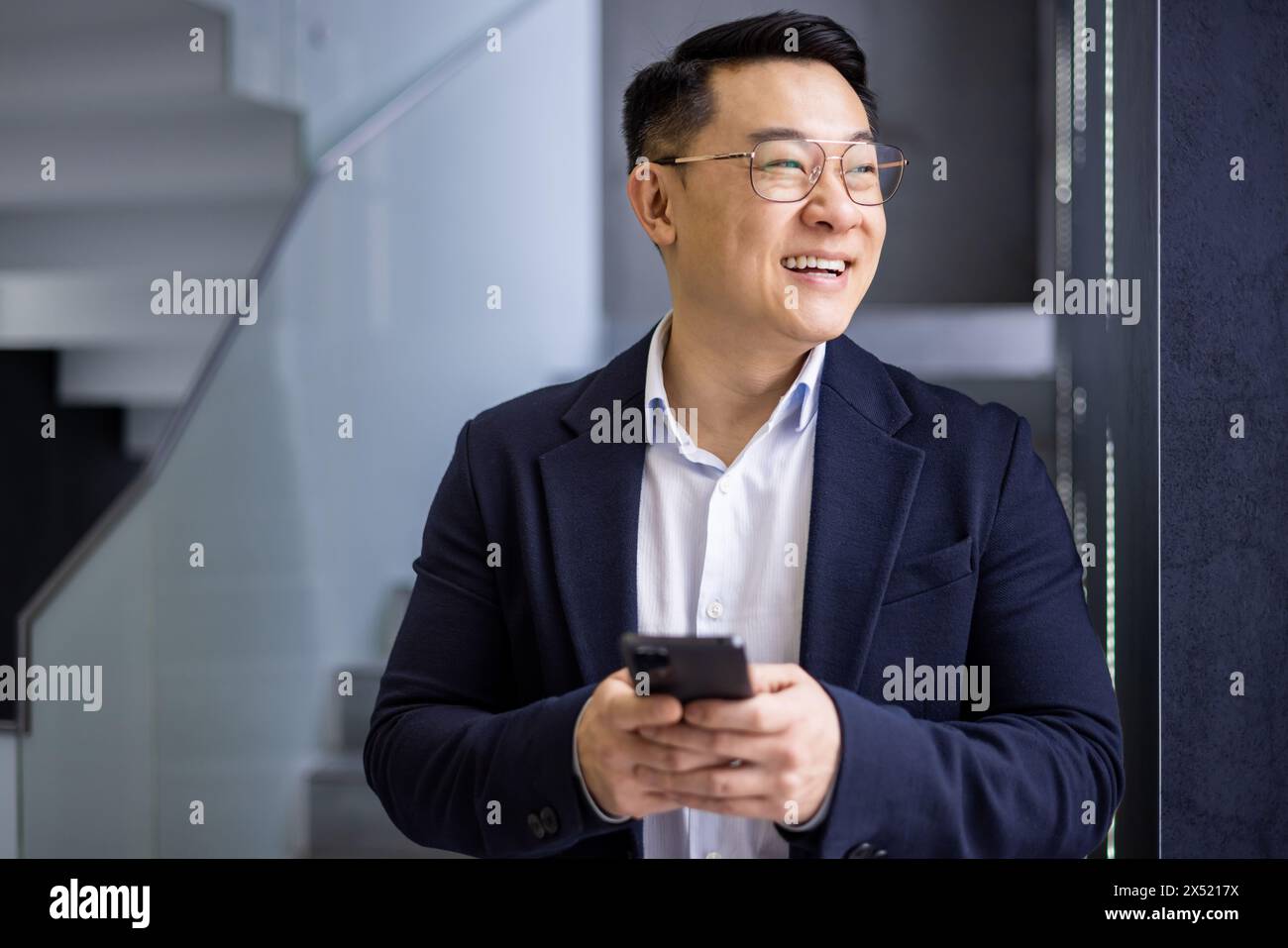 A professional Asian businessman smiling joyfully while using his mobile phone in the contemporary settings of an office staircase, projecting success and connectivity. Stock Photo