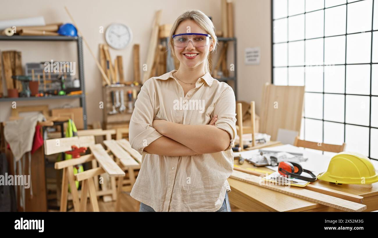 Confident young woman wearing safety glasses stands with arms crossed in a sunny carpentry workshop. Stock Photo