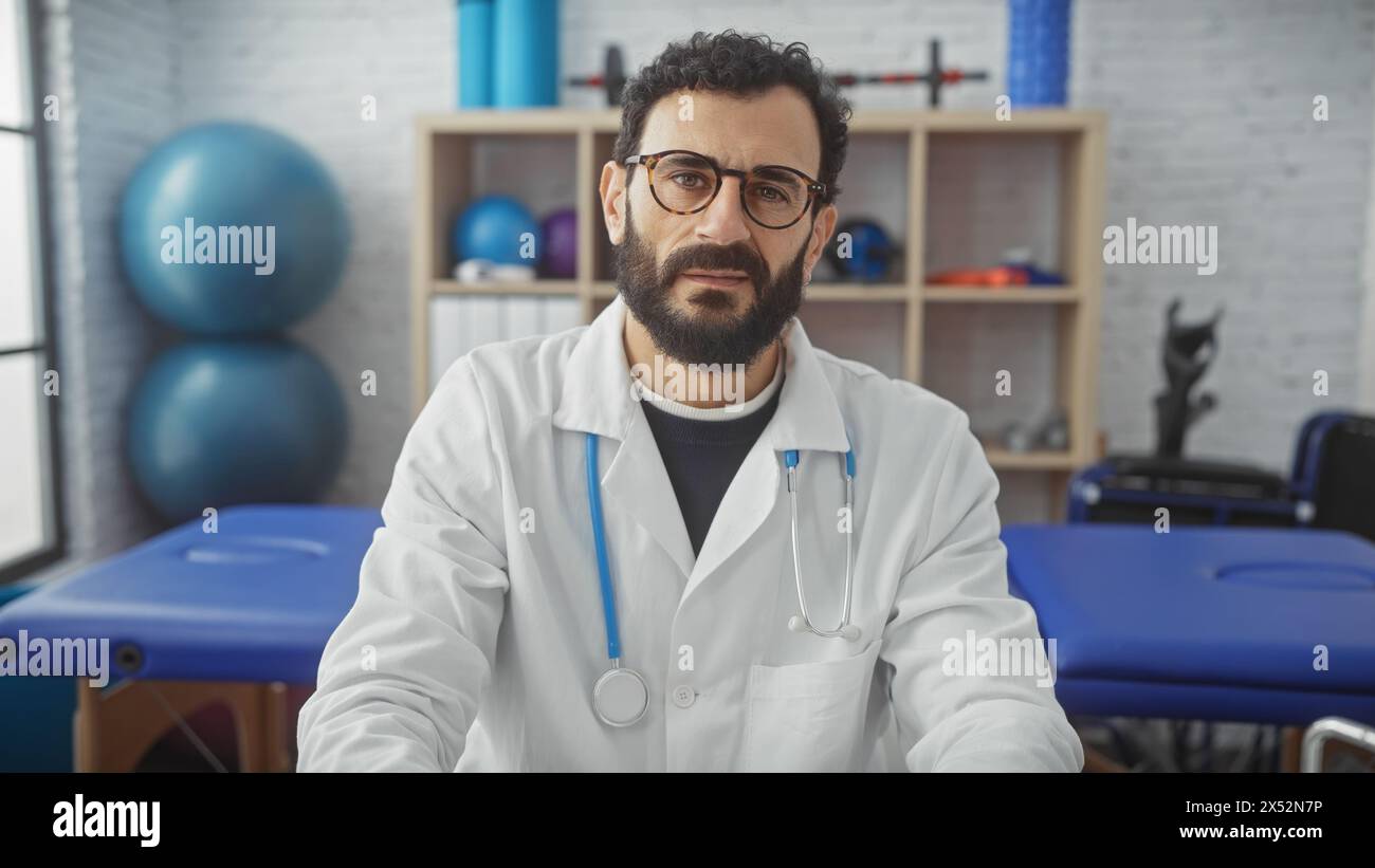 Bearded middle-aged male doctor in a white coat sitting in a rehabilitation clinic's therapy room with exercise equipment in the background. Stock Photo