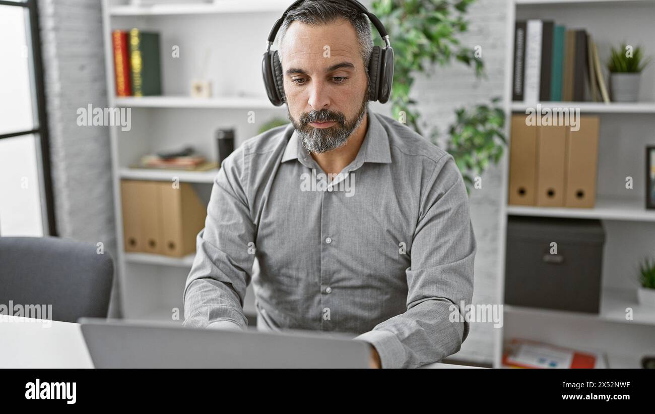 Mature hispanic man with grey beard in headset focusing on laptop in a modern office environment Stock Photo