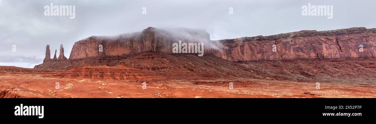 A panorama of the mountain range hosting the famous Three Sisters spires in Monument Valley, Arizona on the left of the image during a cloudy, rainy d Stock Photo